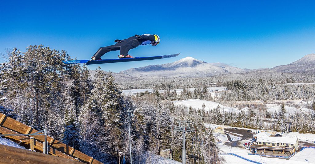 A ski jumper flies through the air during the winter. Its a blue sky day and a snowy Whiteface Mt stands in the background. 