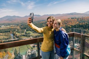 Two young women stand on the observation deck on top of the HS128.They are taking a selfie on a sunny fall day with Adirondacks fall views in the background.